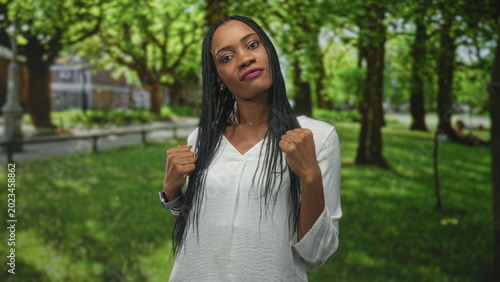 Photos Woman raising fists in forest park wearing white top, bracelets and smartwatch, facing camera with braided hair and assertive pose; confidence resilience