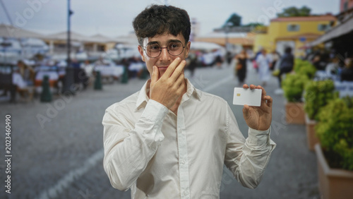 Фотография Young man holding white creditcard with hand covering mouth on street; identity theft concern embarrassment