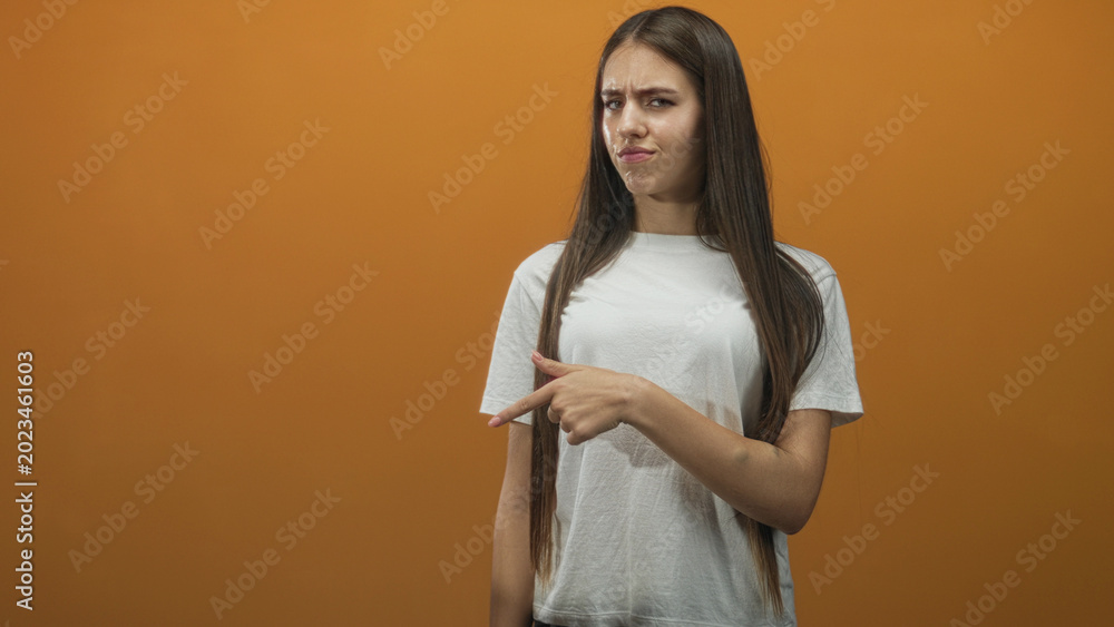 Obraz premium Young woman points finger to right wearing white tshirt in studio with orange backdrop; disapproval.