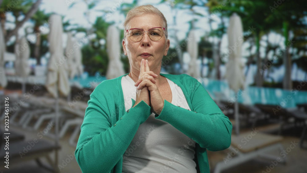 Fototapeta premium Middle aged woman in green cardigan points both hands like gun toward poolside building with sunlit lounge chairs, hands and face in focus; playful defiance.