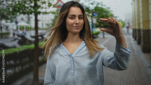 Fotografija Woman in light blue blouse pinching a tiny gap between fingers on a wet city street sidewalk with scooters and trees visible; confidence measurement