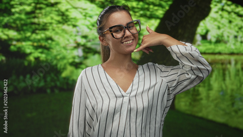 Woman with glasses and striped blouse smiling makes call gesture with hand to face in forest park setting; playful charm.