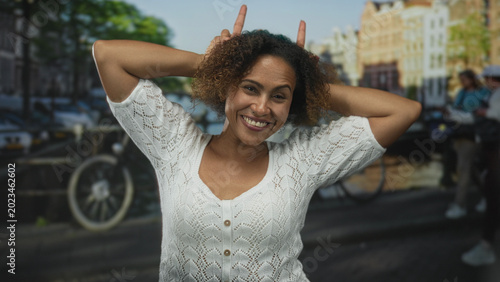 Foto Woman with hands behind head and fingers in hair on an amsterdam street wearing a white cardigan, posing by canal; playful self expression
