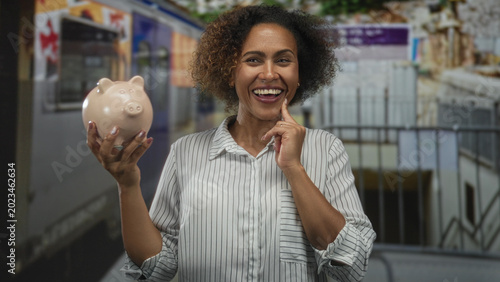 Εκτύπωση καμβά Woman holding piggy bank, finger to cheek, near train in station building, smiling; financial optimism