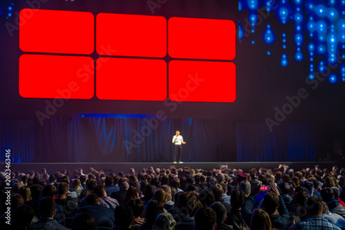 Keynote speaker addresses large crowd at conference, bright red LED panels and blue stage lights create dynamic atmosphere.