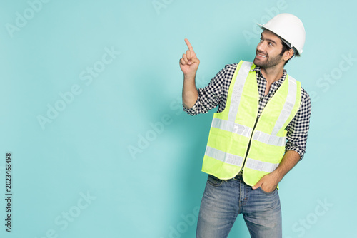 A smiling male engineer in a white hard hat and safety vest points his finger up and looks at copy space, isolated on a cyan background. Great for industrial construction and advertising concepts