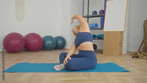 Fotografía Woman seated on blue yoga mat stretching raised arm and holding knee, wearing socks, fitness studio with exercise balls and ropes; calm focus wellness