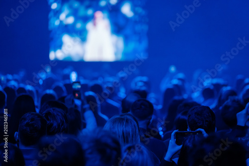 Blue lit concert audience watching blurred stage, crowd using phones to record, energetic atmosphere, shared music experience.