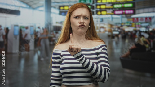 Valokuva Woman points finger with pursed lips and bare shoulder at busy airport gate showing warning gesture; stern disapproval