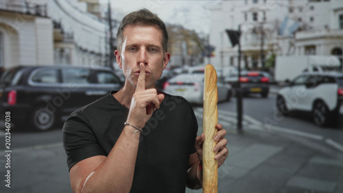 Foto Young caucasian man holding long baguette and finger to lips for silence on busy street in city center; quiet secrecy