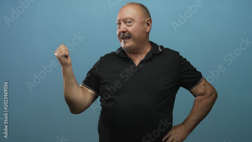 Bild auf Leinwand Senior man smiling with clenched fist raised and hand on hip in studio against blue backdrop; confidence playfulness