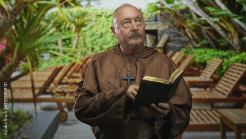 Cuadro en lienzo Senior hispanic monk in brown robe reads gilded bible held in hands on resort deck with wooden lounge chairs; faith contemplation serenity