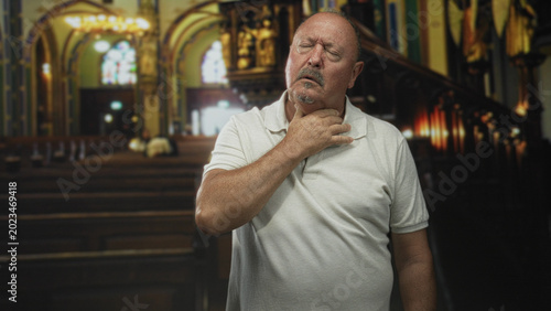 Bild auf Leinwand Man clutching his throat with closed eyes in a church aisle, wearing a white polo and leaning by wooden pews under stained glass light; pain solitude