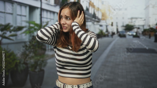 Foto Young redhead woman covers ears with hands on a city street, striped top and studded belt visible; anxiety