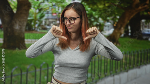 Billede på lærred Young woman with glasses shows both thumbs down, hands and thumb visible, standing by a metal fence and trees on a street; disapproval doubt
