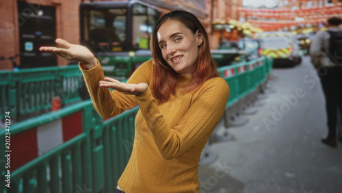 Obraz na plátně Redheaded young woman holds palms up in a presenting gesture by green barriers and a city bus on a busy street; curiosity playful