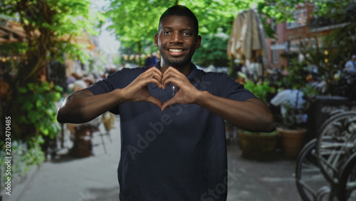 Quadro su tela Man forming heart with hands and smiling on street cafe patio among plants and bicycles; love connection warmth