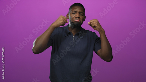 Photography Man raising fists and pouting with closed eyes in studio against a purple backdrop, wearing a polo shirt; playful groove