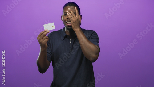 Quadro em tela Man holding creditcard and covering one eye with hand in a purple studio backdrop, mouth open in a startled expression; financial anxiety