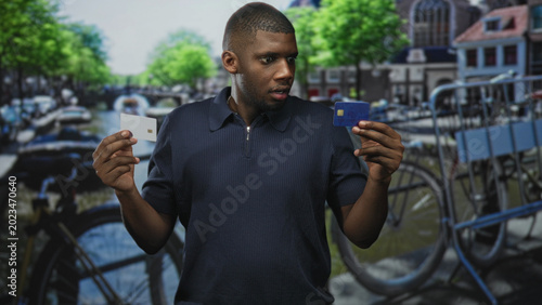 Φωτογραφία Man holding two creditcard, comparing a blue card and a white chip card with a puzzled look on a canal street; doubt decision