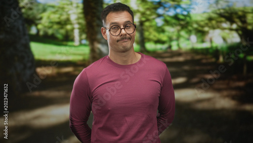 Man with glasses and mustache smirks, head tilted and hands behind back in a sunlit park wearing a magenta long sleeve shirt; casual amusement.