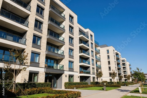 Modern apartment buildings exterior with balconies under clear blue sky. Contemporary architecture features light brick and dark accents. Landscaped green areas surround residential units.