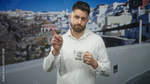 Fotografie Man holding coffee mug that reads i am the boss, pointing finger with right hand in studio set, white shirt and beard; irony leadership