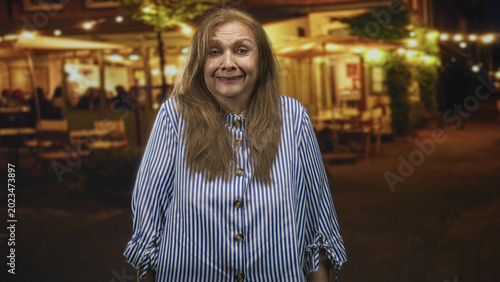 Konstfotografi Woman smiling with visible face and striped blouse standing outside a restaurant on a street at night; contentment evening warmth