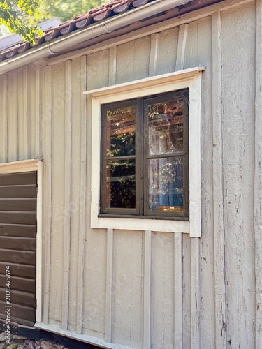 Traditional dark wooden farmhouse window with soft reflections of surrounding trees and sunlight. Rustic Scandinavian countryside architecture with nostalgic rural mood.