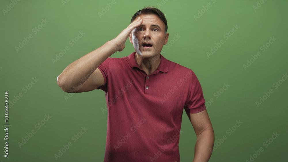 Fototapeta premium Man touching forehead with hand in studio green backdrop, wearing maroon polo and standing midshot; frustration.