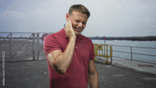 Принти на полотні Handsome hispanic man with hand on cheek wincing near pier railing and yellow barrier by the water; toothache pain