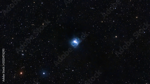 Observe a brilliant blue reflection nebula luminous against the dark cosmic expanse. Numerous white and orange stars twinkle across the deep black sky.
