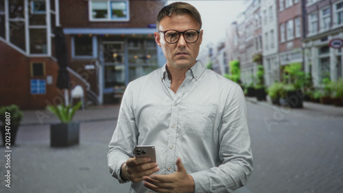 Foto Man holds smartphone and gestures with hands while frowning and checking screen on urban street, wearing glasses and casual shirt; frustration technology