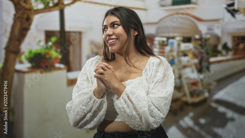 Photos Woman in a white embroidered blouse clasping hands near chest and smiling on a mediterranean street beside a souvenir kiosk and planter; gentle joy
