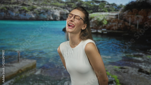 Fotografie Woman in white sleeveless top smiling with tongue out and bare shoulders visible beside a rocky coastal pier at the seaside; playful summer escape