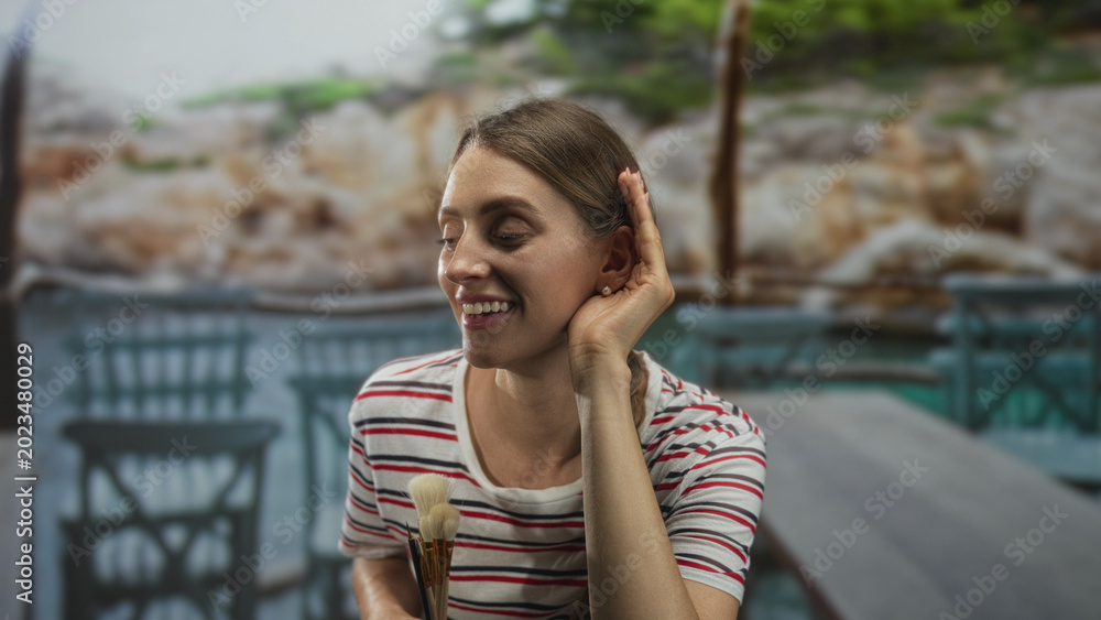 Fototapeta premium Woman holding paintbrushes cups ear with hand while smiling at a seaside cafe table in studio setting; serene creativity.
