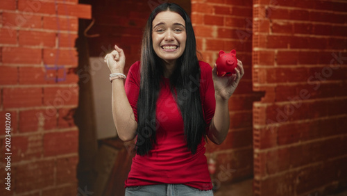 фотографія Woman smiling and holding a red piggybank with raised hand against exposed brick wall in studio; saving optimism security