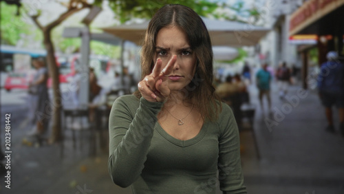 Canvastavla Young hispanic woman points two fingers to nose at street cafe terrace outdoors while seated at a table, wearing a heart pendant; annoyance impatience