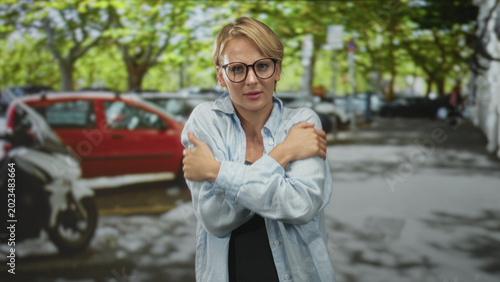 Blond woman hugging her shoulders with crossed arms on a leafy street beside parked cars and a scooter under tree shade; cold.