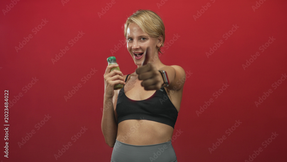 Naklejka premium Woman with blond hair in black sports bra holds green juice bottle, smiles, points forward and shows thumbs up in studio; confidence vitality.