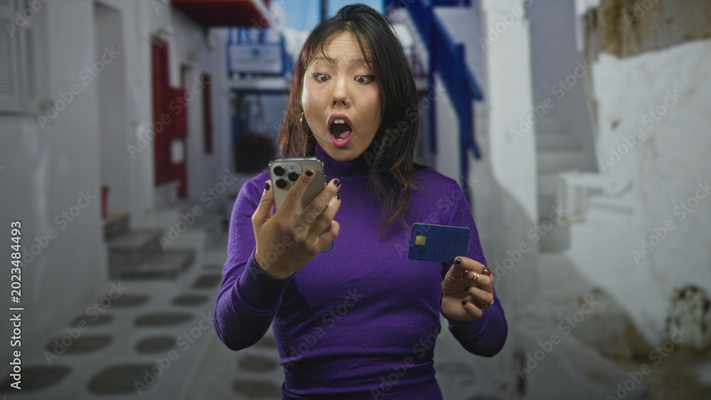 Fototapeta premium Young chinese woman holding smartphone and creditcard, mouth open in surprise on narrow white street; surprise discovery.