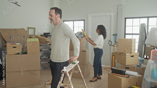Photos Man and woman pack stacked cardboard boxes and seal tape while man holds a ladder and woman uses a tape dispenser, moving supplies and dog in building; hopeful new beginning
