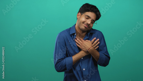 Bilde på lerret Man with hands on chest, wearing blue shirt and glasses, eyes closed and smiling against a green backdrop in a studio; gratitude sincerity warmth