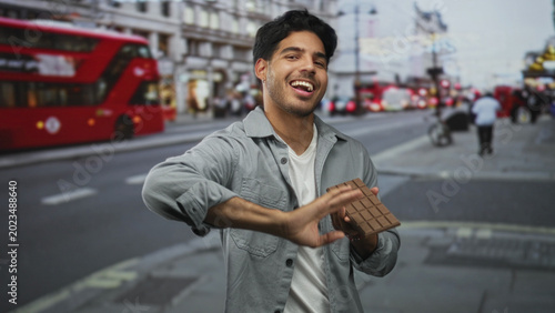 Photography Man holding chocolate bar in his hand smiling on a london street with a red doubledecker bus and blurred pedestrians; joy snack casual