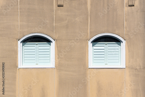 Two white shuttered windows frame a textured beige wall. Simple architecture adds calm, minimalist aesthetic. Clean lines contrast subtle wall grain. Bright, even light enhances window shapes
