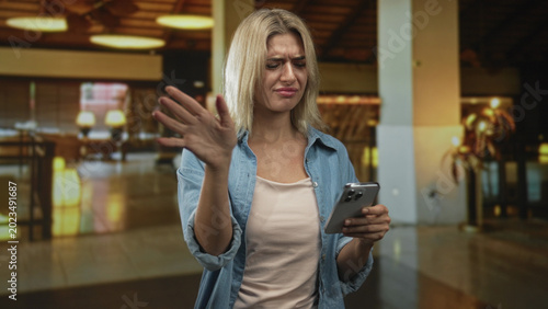 Fotografie Woman frowning while holding smartphone and checking messages in building lobby with lamps and seating; frustration communication