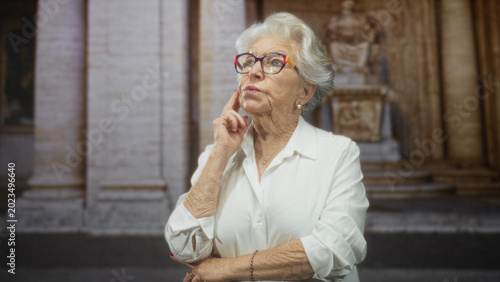 Foto Senior woman wearing colorful glasses touches cheek with index finger and crosses arm in front of classical stone building with columns; thoughtful reflection