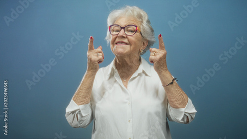 Tableau sur toile Senior woman gestures with index fingers to her cheeks then points both index fingers upward, wearing colorful glasses and white blouse in studio; playful joy