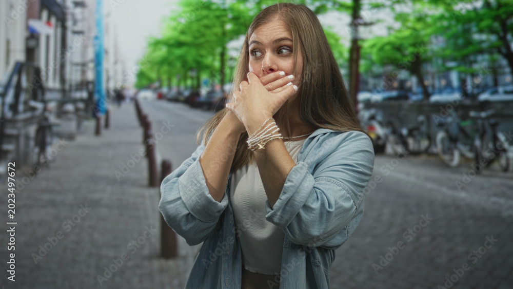 Fototapeta premium Young blonde woman covers mouth with both hands against lips on busy city street in daylight; hush reflection.