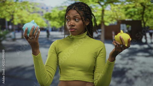 Fotografi Woman holding blue and yellow piggy banks on street with trees and sidewalk while comparing savings options; money choices contemplation